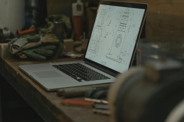 Laptop showing a technical drawing, surrounded by tools and gloves in a workshop setting.